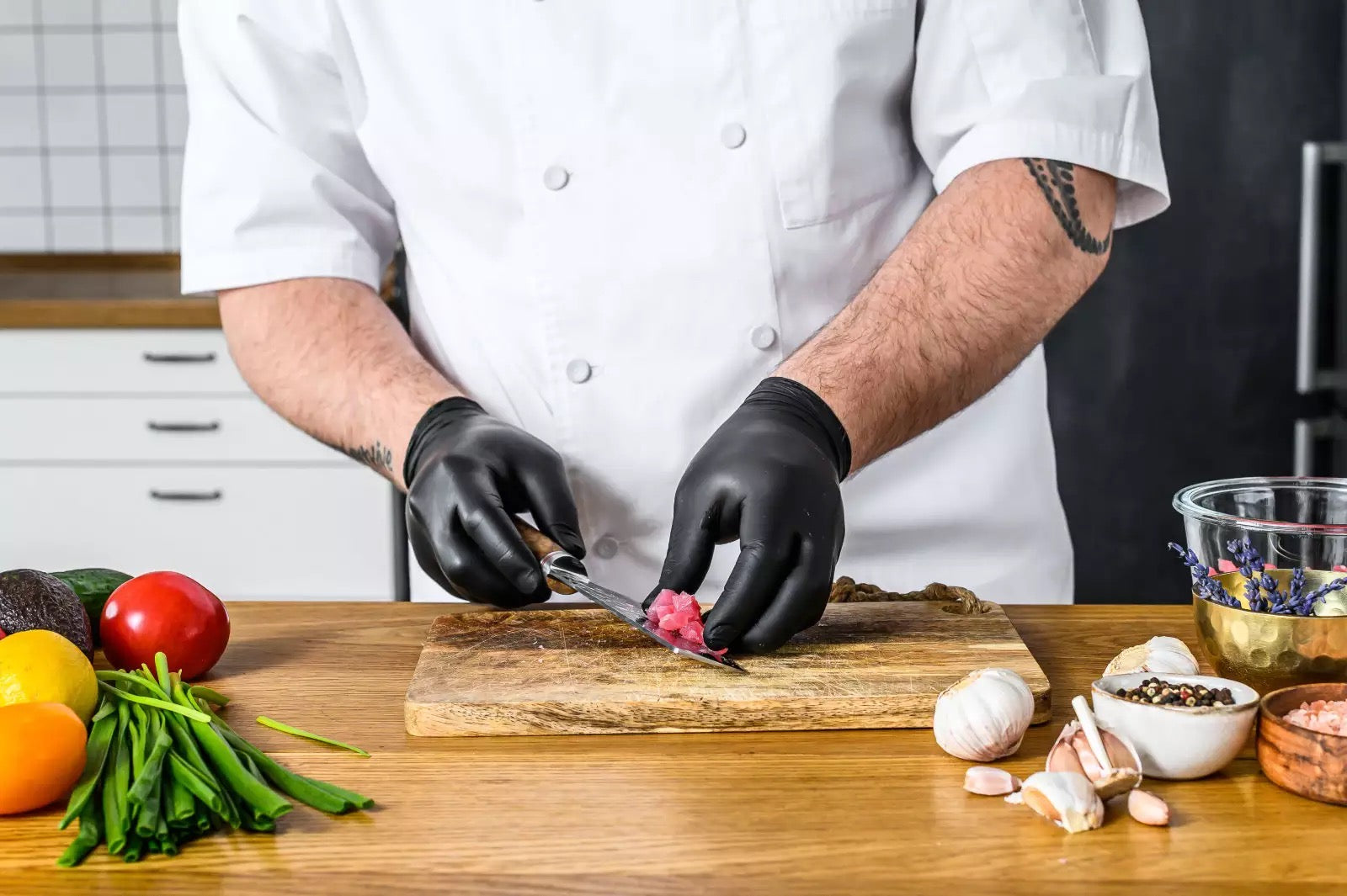 Chef in a kitchen wearing a black nitrile glove preparing food on a wooden cutting board with various vegetables and ingredients.