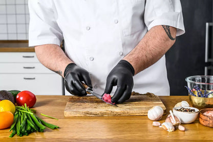 Chef in a kitchen wearing a black nitrile glove preparing food on a wooden cutting board with various vegetables and ingredients.