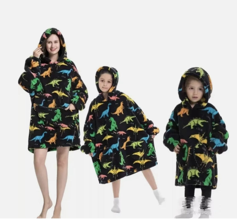 Three children wearing colorful dinosaur-patterned oodie blancket hoodies on a white background